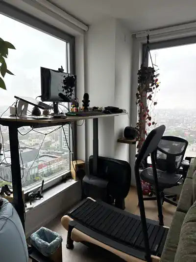 A standing desk setup by a large window with a city view, featuring a monitor, keyboard, microphone, office chair, treadmill, and various cables and accessories. A plant and fairy lights decorate the corner.