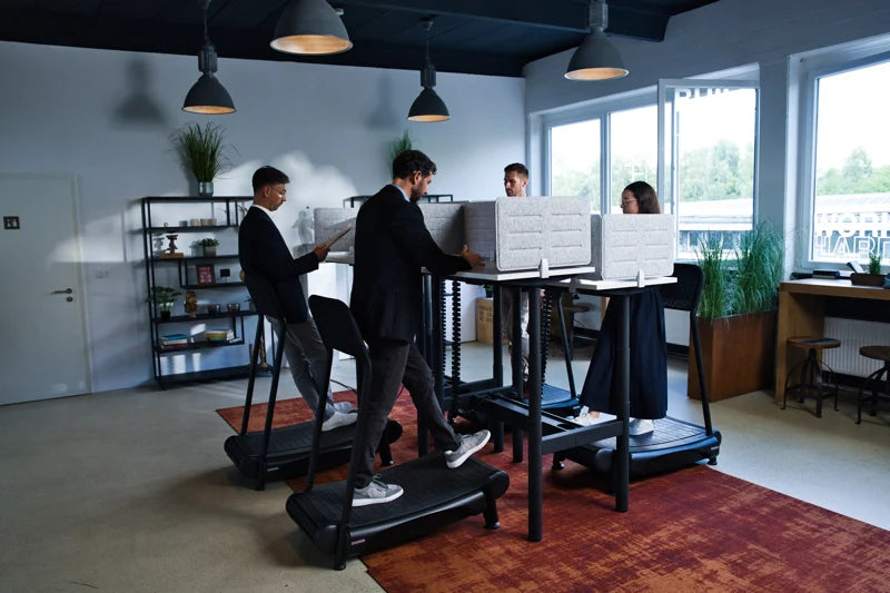 Four people work at standing desks with treadmills in a modern, bright office. Shelves with books and plants are in the background, and large windows let in natural light. Walkolution Global
