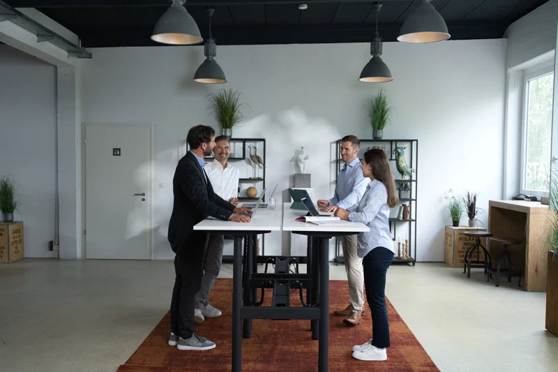 Four people stand around a high table in a modern office with shelves, plants, and laptops. They appear to be having a meeting or discussion in a bright, well-lit room. Walkolution Global
