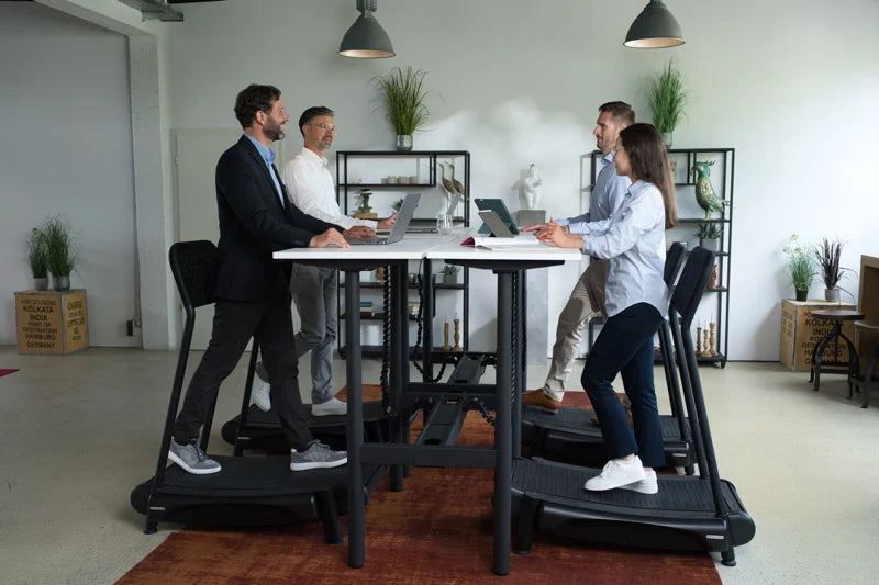 Four people work on laptops at a tall shared desk, each standing on individual treadmill desks in a modern office with plants and shelves in the background. Walkolution Global