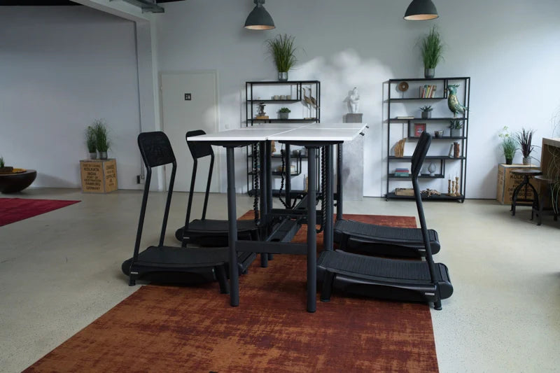 A modern office space with a rectangular table surrounded by four treadmill desks, each with a black chair. Shelves with plants and decor are in the background, and the floor has a large reddish-brown rug. Walkolution Global