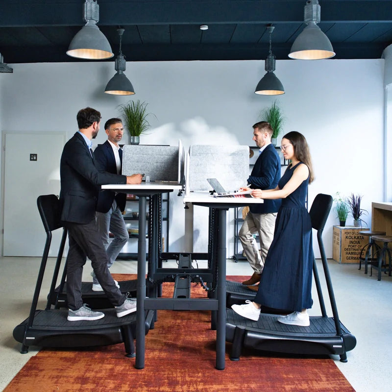 Four people are working at standing desks with treadmill bases in a modern office. Two men and one woman use laptops, while another man stands. The desks have privacy screens, and pendant lights hang from the ceiling.