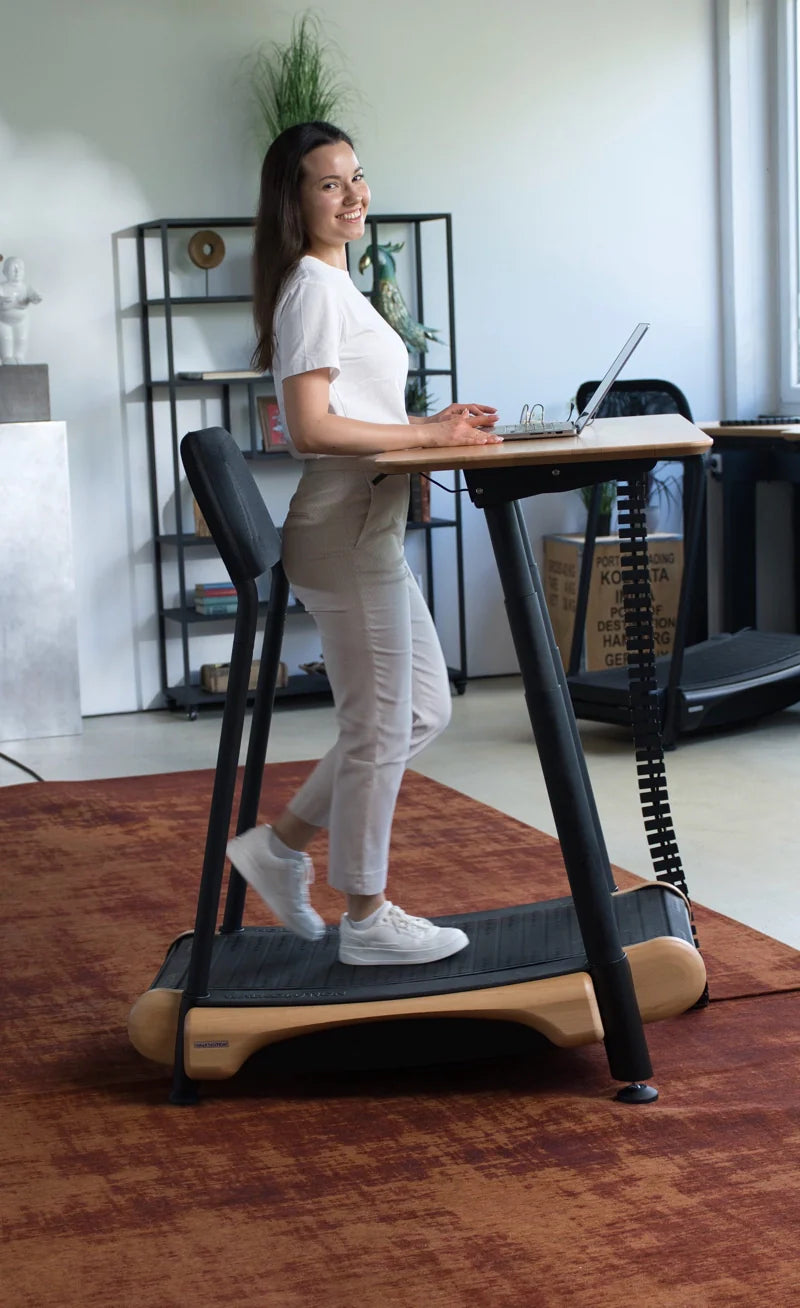 A woman smiles while walking on a treadmill desk and working on a laptop in a modern office space with shelves and a large window. Walkolution Global