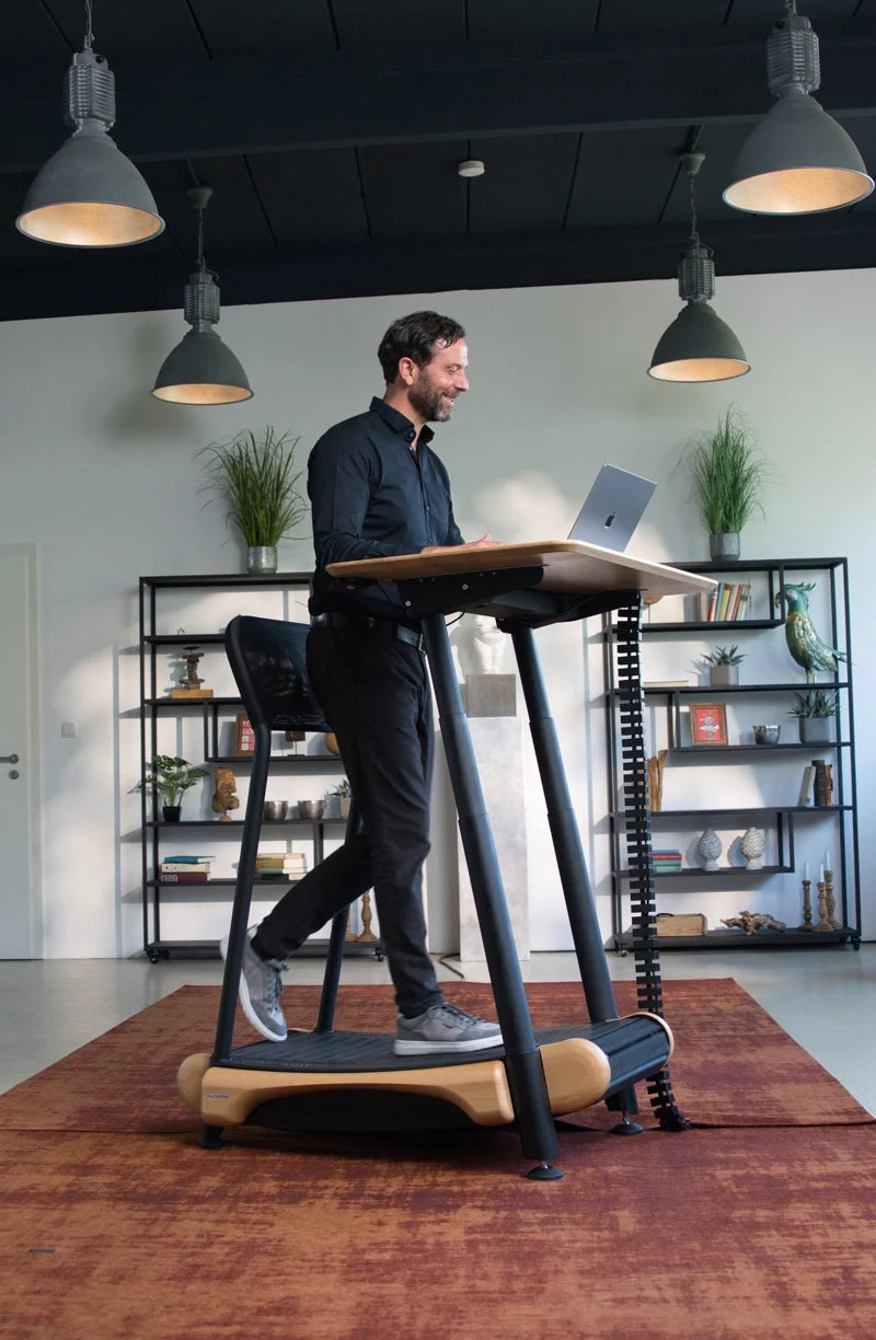 A man in dark clothing works on a laptop at a standing desk treadmill in a modern, well-lit office with plants, shelves, and decorative items in the background. Walkolution Global