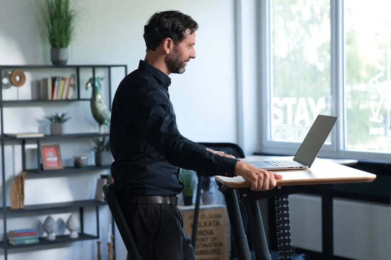 A man in a black shirt stands at a height-adjustable desk, working on a laptop in a modern office with shelves, books, and a large window. Walkolution Global