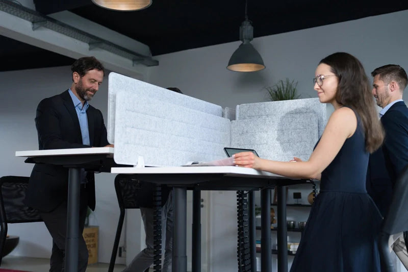 Four people working at height-adjustable desks with privacy partitions in a modern office. One woman is seated with a tablet, while three men are standing and engaged in conversation. The space has industrial lighting.