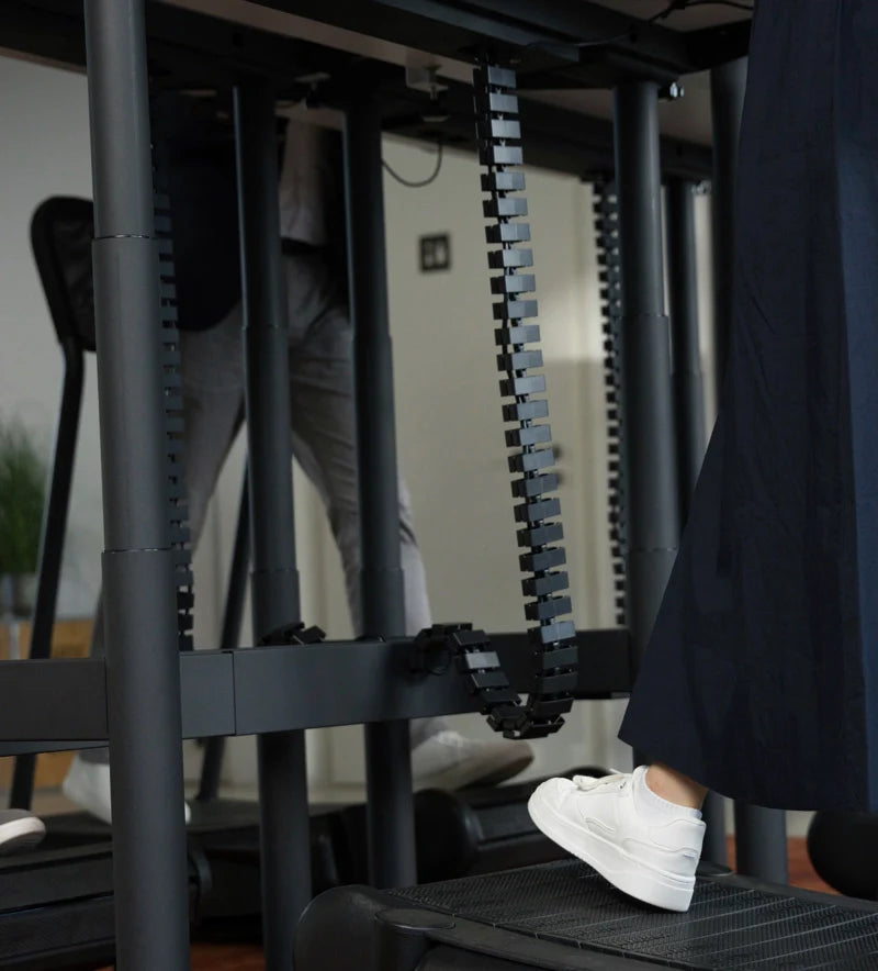 Close-up of two people walking on treadmills under the height-adjustable Walkolution Forum Conference Desk. One wears white sneakers and a dark skirt, the other light pants. Visible cable management links the desk and treadmills.