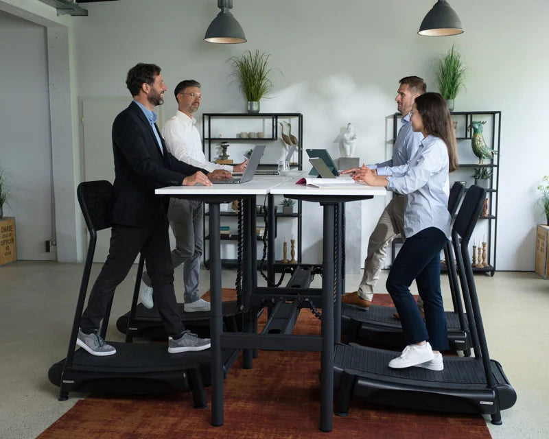 Four people in business attire work on laptops at a tall shared desk, each standing on a treadmill. The modern office background includes shelves with plants and decor, under hanging lights.