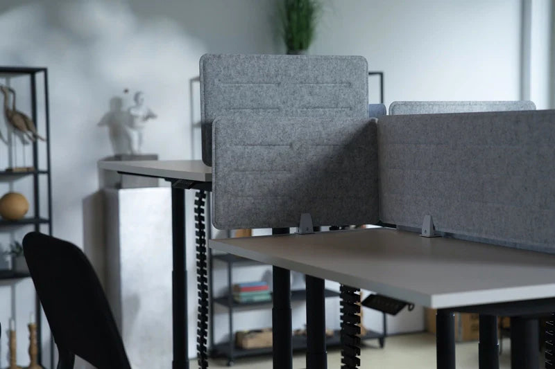 A modern office features empty Walkolution Forum Conference Desks separated by gray privacy panels, paired with black chairs and shelves in the background displaying decorative items and plants.