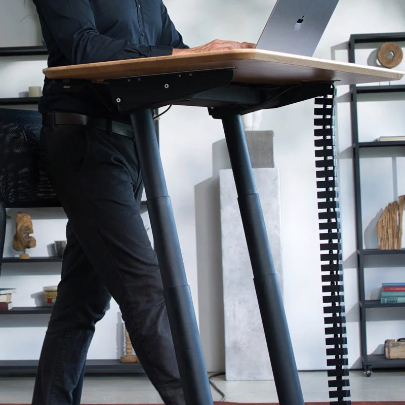 A person works on a laptop at a Walkolution Fusion Desk, an electric height adjustable desk, creating an ergonomic workspace in a modern office with shelves and books in the background.