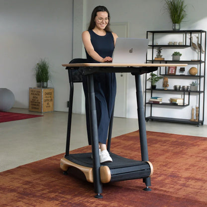 A woman works on her laptop at a Walkolution Fusion Desk treadmill workstation in a modern office with ergonomic features, shelves, plants, and a red rug in the background.