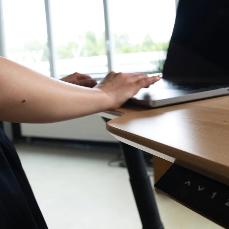 A person uses a laptop on the Walkolution Fusion Desk, an electric height adjustable desk with ergonomic features, positioned near a window overlooking blurred greenery.