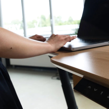 A person uses a laptop on the Walkolution Fusion Desk, an electric height adjustable desk with ergonomic features, positioned near a window overlooking blurred greenery.