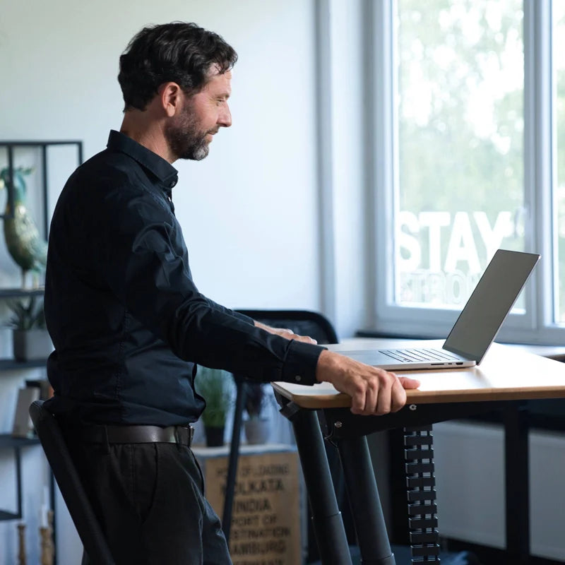 A man in a black shirt works on a laptop at the Walkolution Fusion Desk, an electric height adjustable desk, in a bright, modern room with large windows, creating an ergonomic workspace.