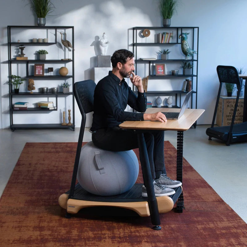 A man works on a laptop, seated on a gray exercise ball at a Walkolution Fusion Desk, with shelves, plants, and decorative objects in the modern office background.