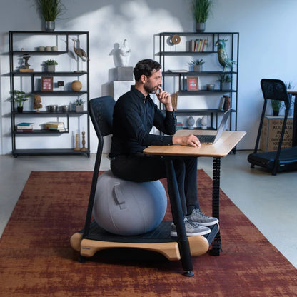 A man works on a laptop, seated on a gray exercise ball at a Walkolution Fusion Desk, with shelves, plants, and decorative objects in the modern office background.