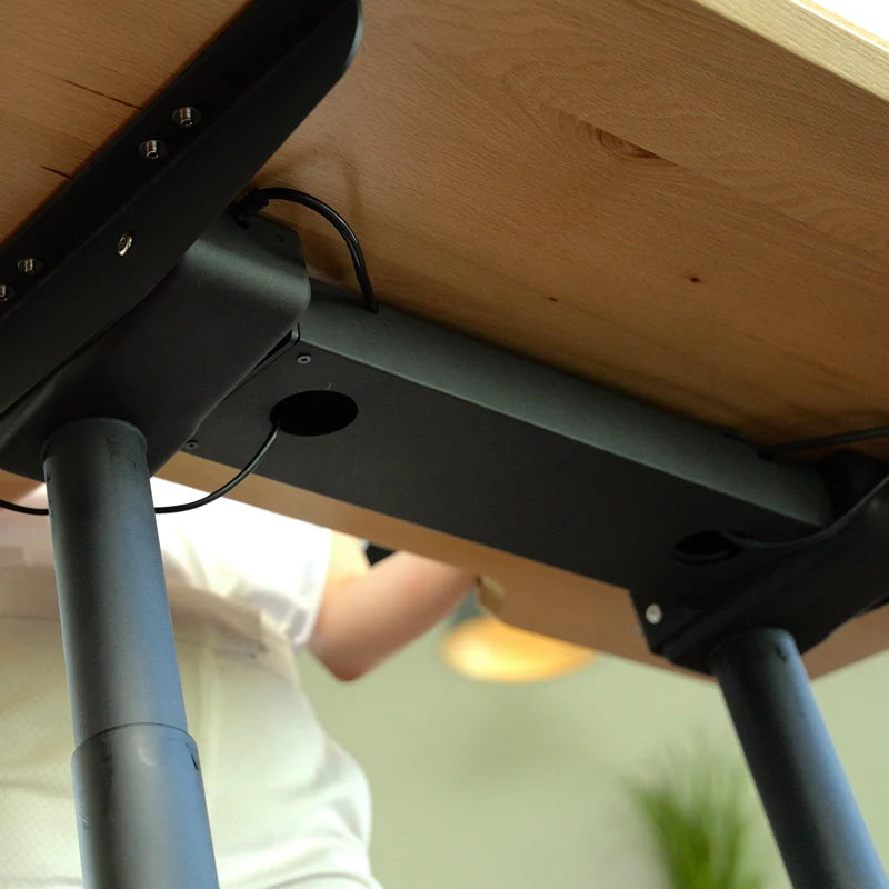 View from under a Walkolution Fusion Desk showing black metal legs and cables in a management tray. A person in light clothing is partially visible, with blurred lighting and a plant, highlighting an ergonomic workspace.