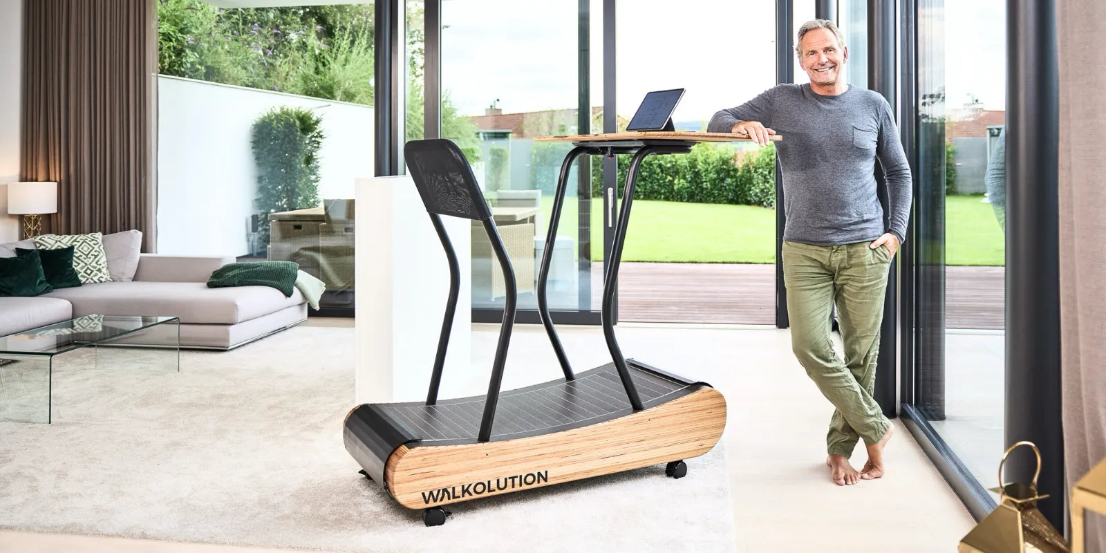 A man stands barefoot, smiling next to a wooden WALKOLUTION treadmill desk with a laptop. He is in a modern, bright living room with large windows and a view of a green yard outside.