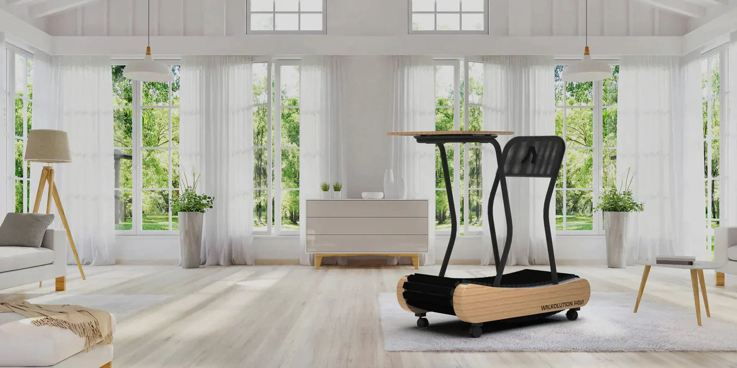 A modern treadmill with wood accents and a desktop stands in a bright, spacious room with large windows, white walls, and minimal furniture, overlooking a green garden.