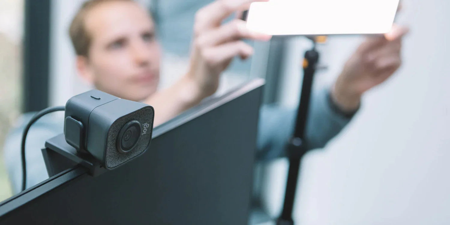 A person adjusts a bright light panel in front of a monitor with a webcam mounted on top, preparing for a video call or recording in a bright indoor setting.