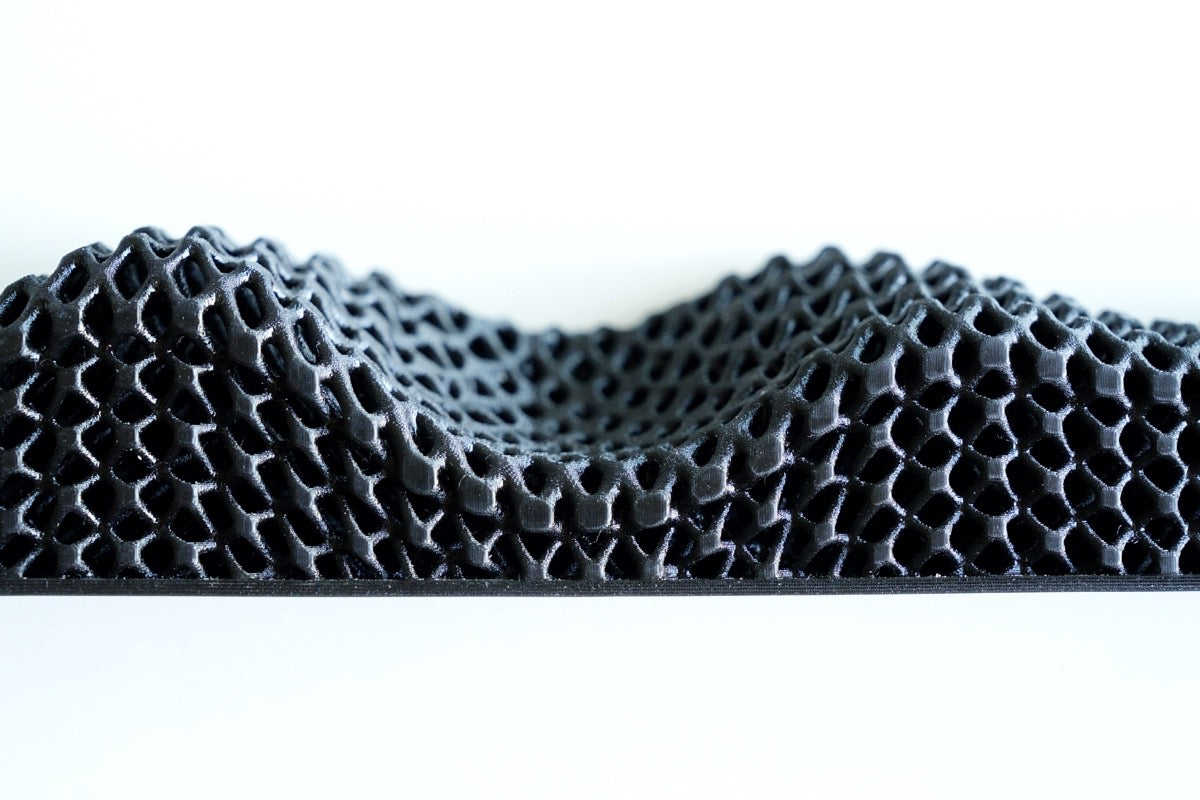 A close-up of a black, 3D-printed honeycomb structure from the All Surfaces Sample Box, featuring wave-like peaks and valleys for barefoot comfort, viewed from the side against a white background.
