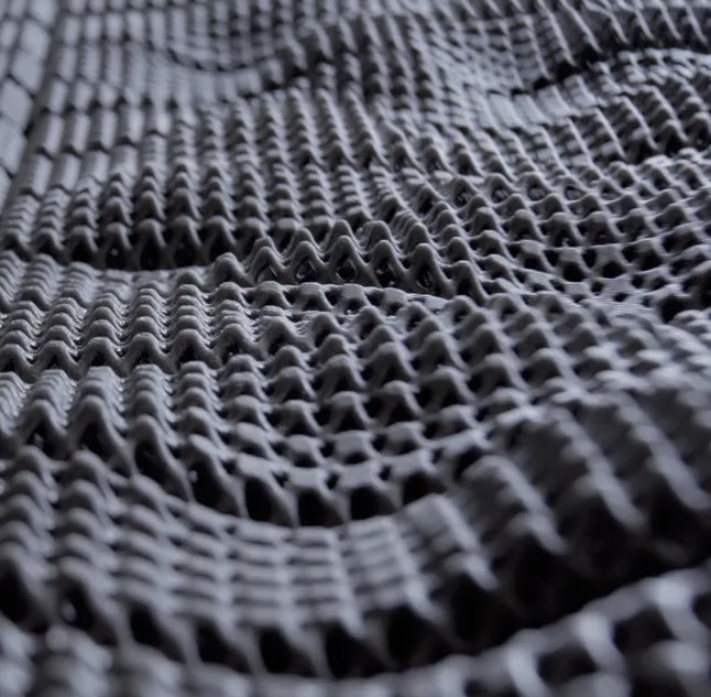 Close-up of a textured black mesh fabric with a wavy, three-dimensional pattern and small, evenly spaced holes, creating a sense of depth and movement across the surface.