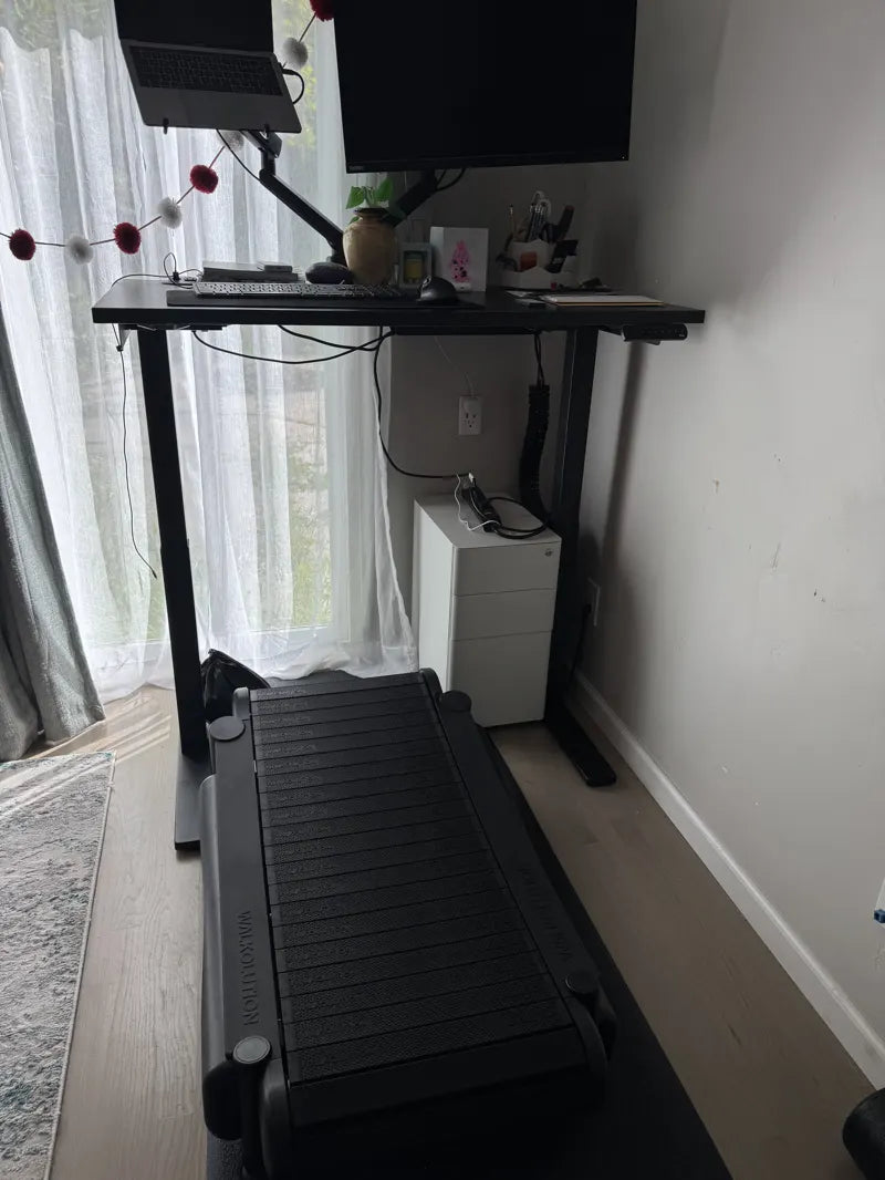 A modern home office setup with a standing desk, computer monitor, laptop, and a treadmill placed in front of the desk. Natural light comes through sheer curtains, and the space includes plants and organized accessories.