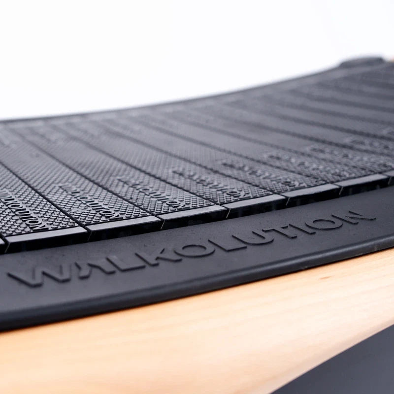 Close-up of a curved black treadmill belt with textured slats and the word WALKOLUTION embossed on the side, set against a light wood and white background.