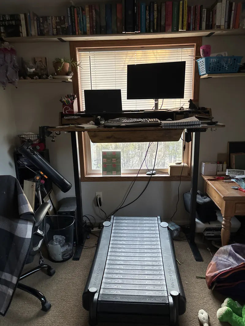 A treadmill desk setup in front of a window, with two monitors, a keyboard, and a laptop on a raised desk. Books line the shelf above, and cluttered furniture and items fill the surrounding room.