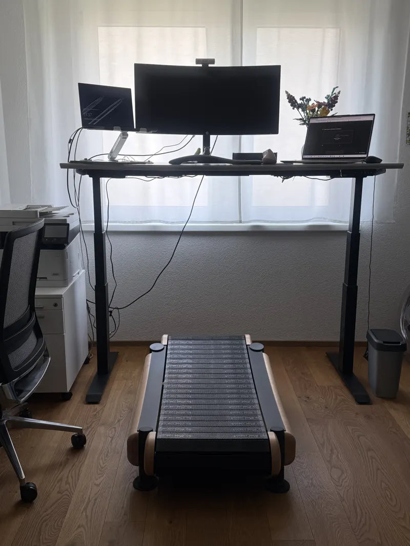 A standing desk setup with two monitors, a laptop, office chair, and a small treadmill under the desk. The workspace is by a window with white curtains and a vase of flowers. Cables and office equipment are visible.