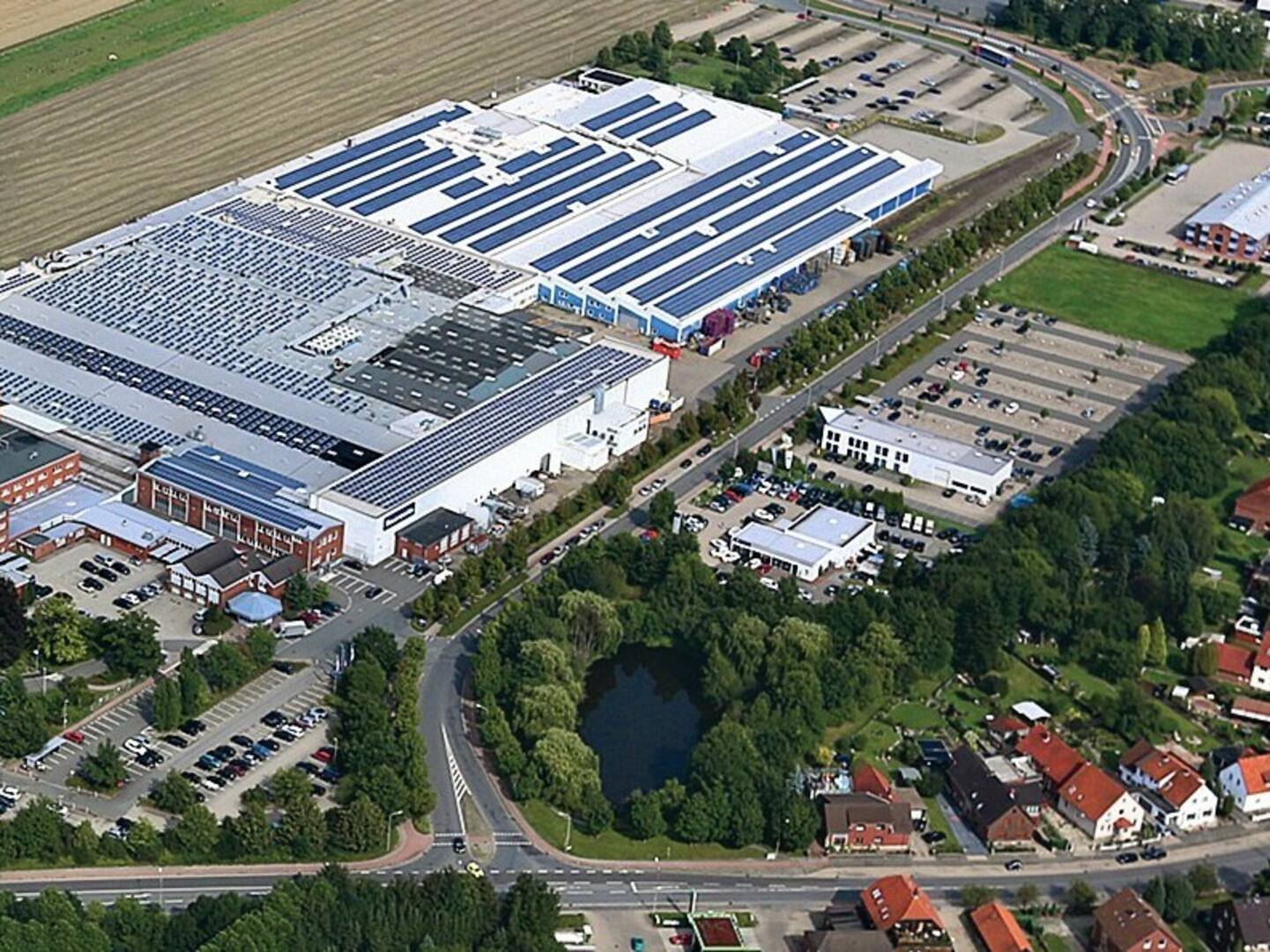 Aerial view of a large industrial facility with solar panels on the roof, surrounded by parking lots, greenery, a small pond, and residential houses nearby.