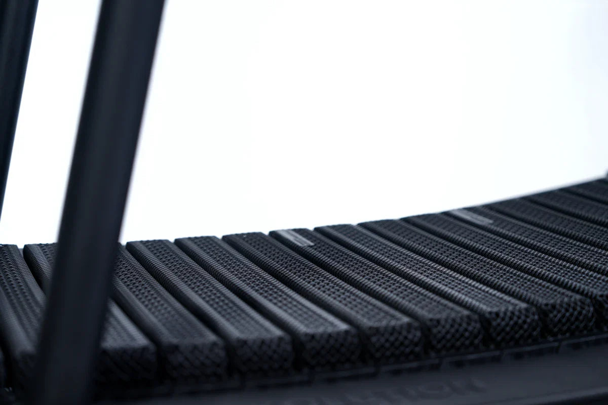 Close-up of the textured black running belt of a curved treadmill, showing its individual slats and part of the treadmill’s frame against a plain light background.