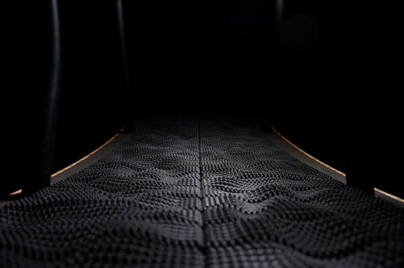 A close-up view of a textured, wavy black rubber surface, possibly a mat or flooring, with soft lighting highlighting the raised patterns. The background is mostly dark, emphasizing the surface details.