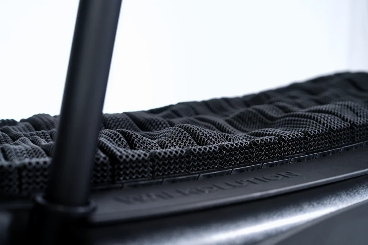 Close-up view of a textured black mesh seat cushion on a chair, with visible grooves and contoured surface, supported by a sturdy black frame.