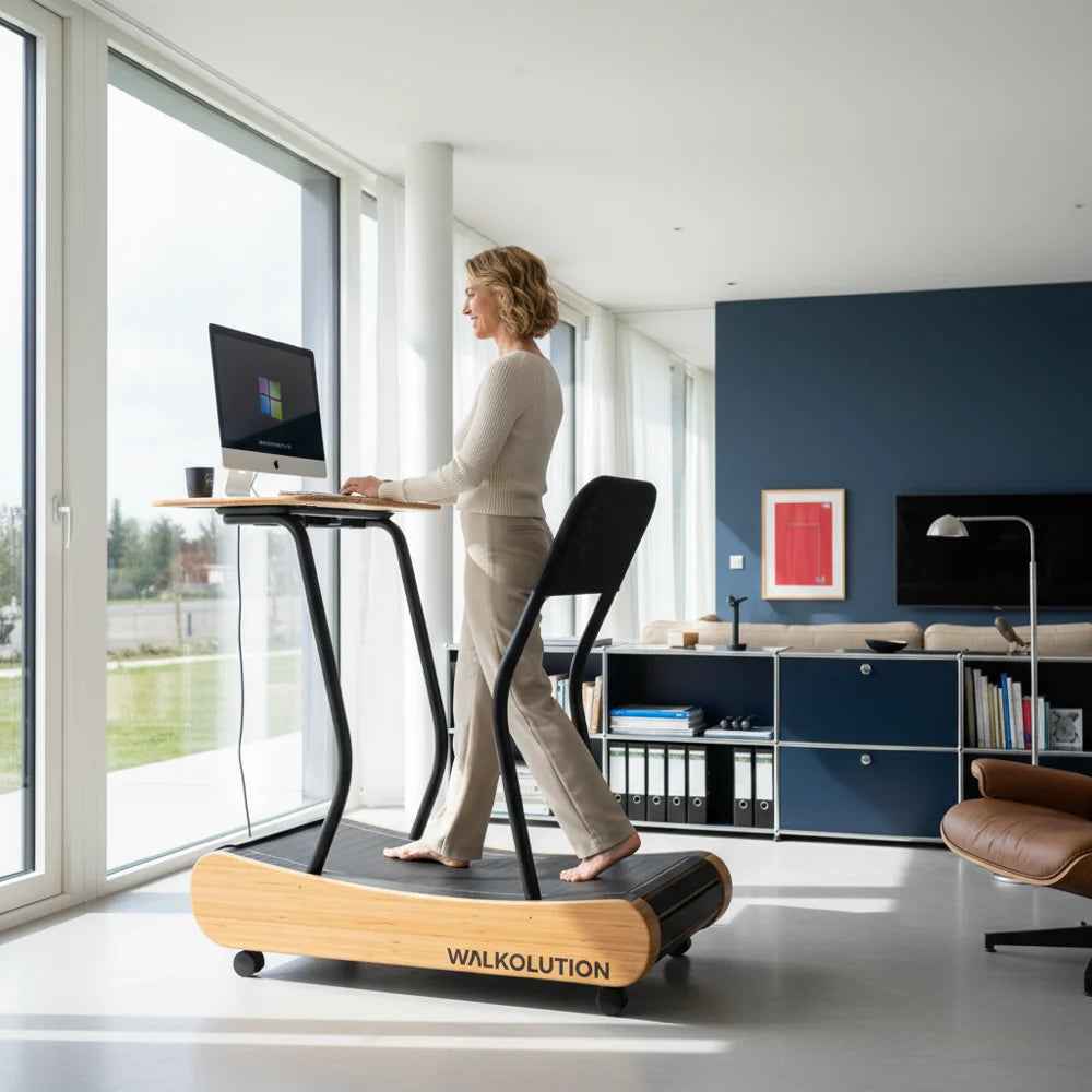 A woman works at a standing desk with a computer while walking barefoot on a wooden treadmill in a bright, modern living room with large windows.