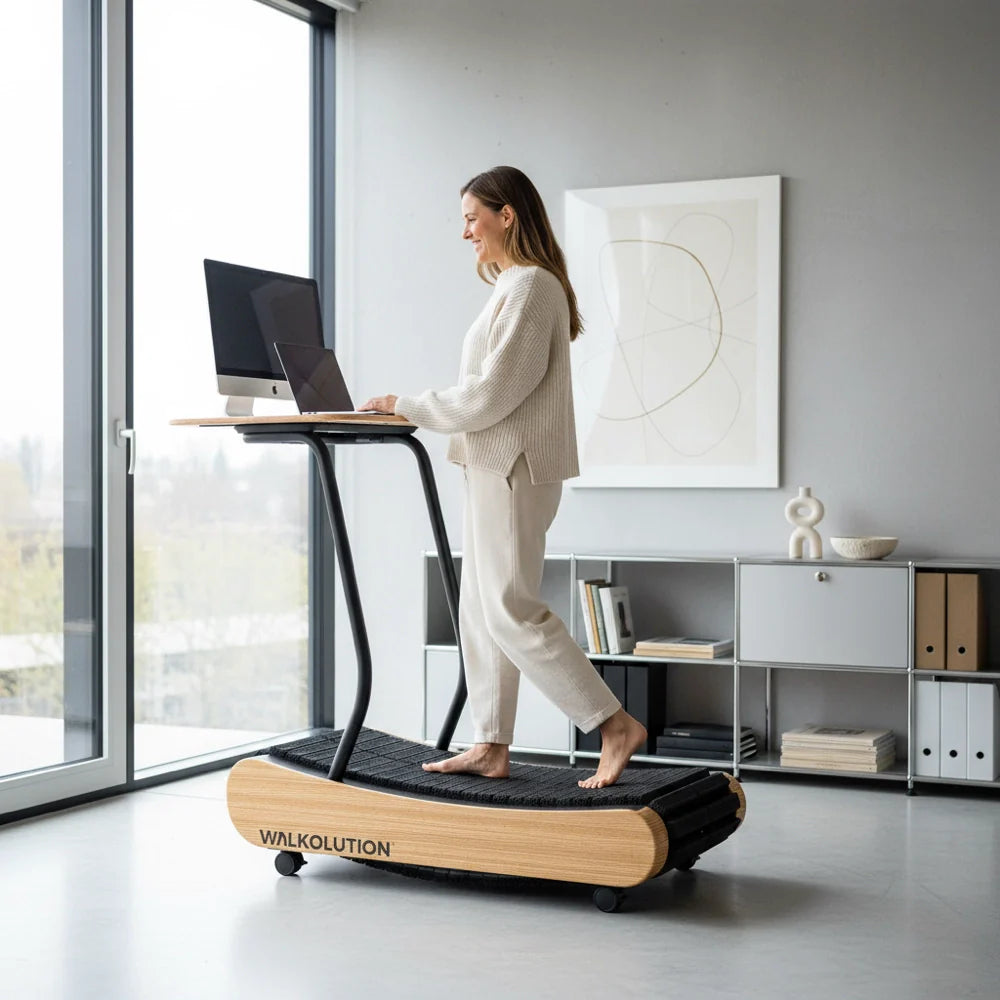 A woman in light-colored clothes walks barefoot on a wooden treadmill desk, working on a laptop in a modern, bright room with large windows and minimalist decor.