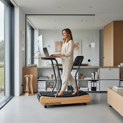 A woman in light loungewear uses the Walkolution 1 manual treadmill as she works at her computer in a bright, minimalist home office with large windows, maximizing her walking desk setup.