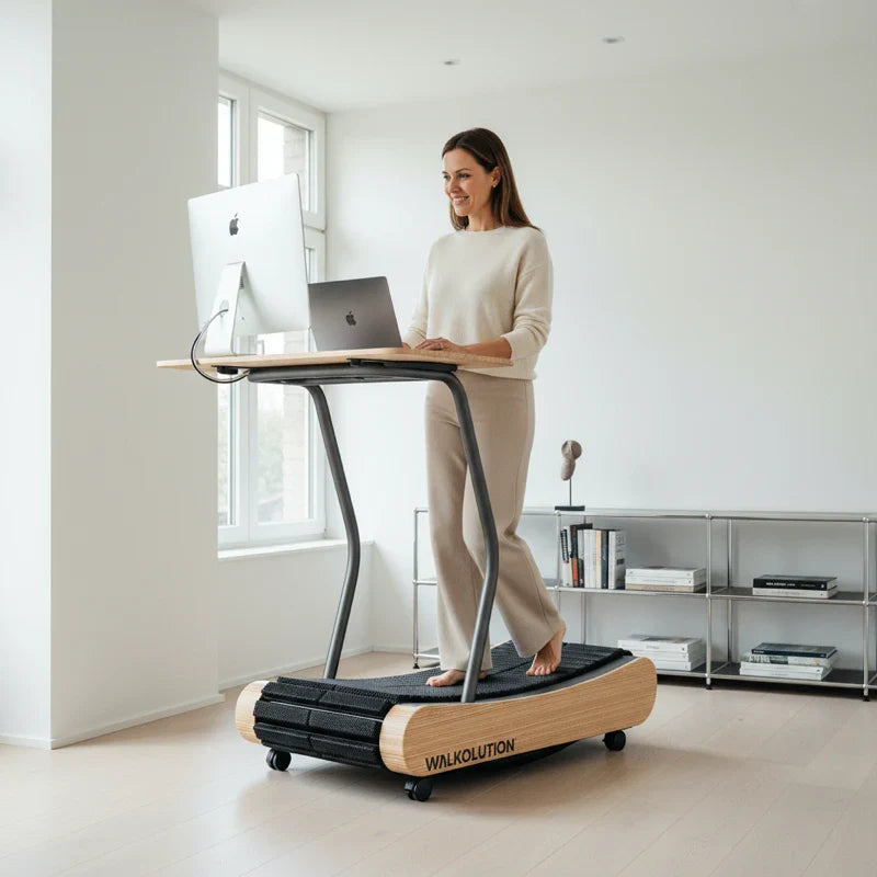 A woman stands barefoot on a wooden treadmill desk with the Walkolution 1 Desk Attachment, working on her laptop in a bright, modern room with minimalist decor and large windows.