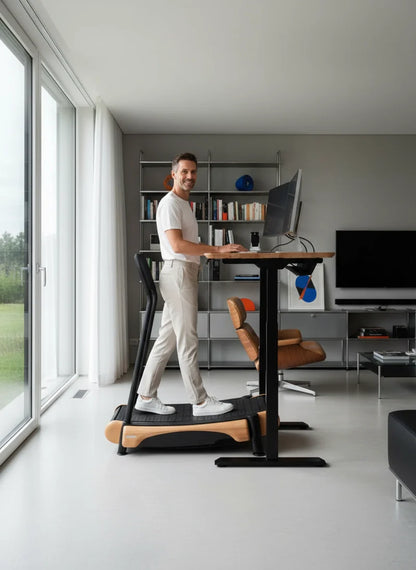A man in casual clothes uses the Walkolution 2 manual office treadmill while working on his computer in a modern home office with large windows, a bookshelf, and minimalist decor for a productive atmosphere.