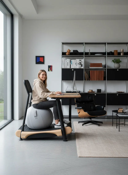 A woman works on her laptop at a Walkolution Fusion Desk in a bright, minimalist home office with shelves, books, and decor. She sits on an exercise ball chair, while large windows fill the space with natural light.