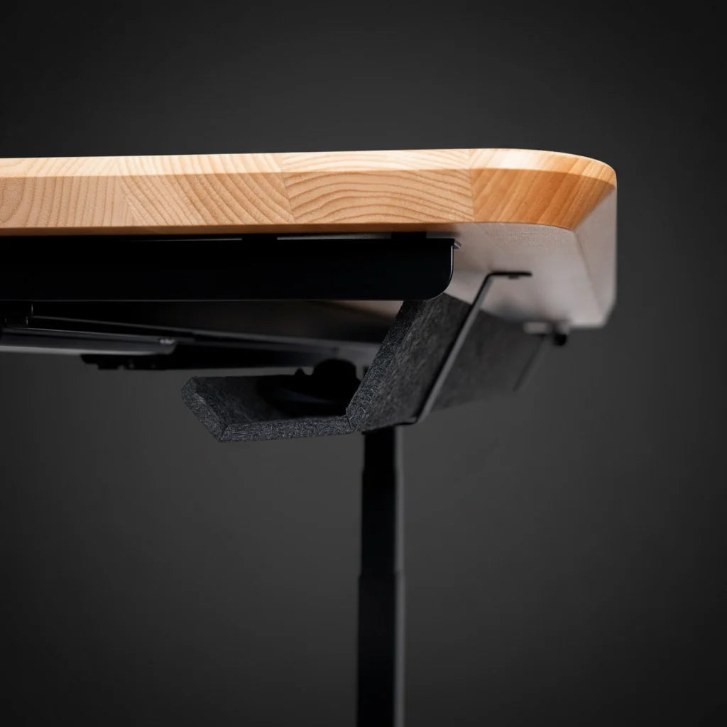 Close-up of the edge of a wooden tabletop with a smooth, rounded finish, supported by a black metal frame and an attached black felt accessory underneath, set against a dark background. Walkolution Global