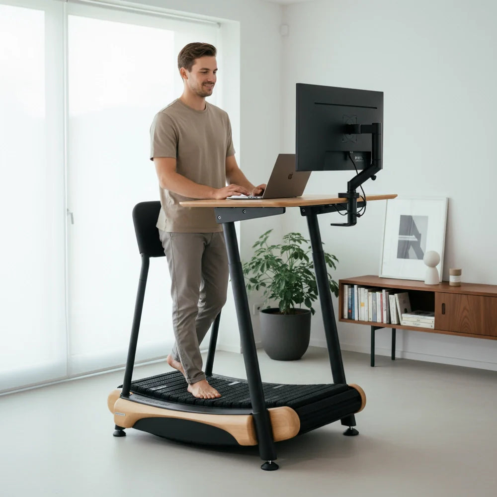 A man stands barefoot on a treadmill desk while working on a computer in a modern, minimalistic home office with a plant and cabinet in the background.