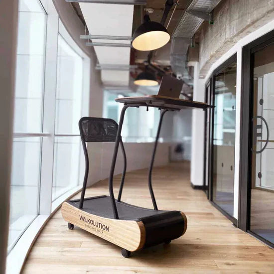 A treadmill desk with a wooden base and a raised workstation for a laptop is set up in a modern office hallway with large windows and wooden floors.