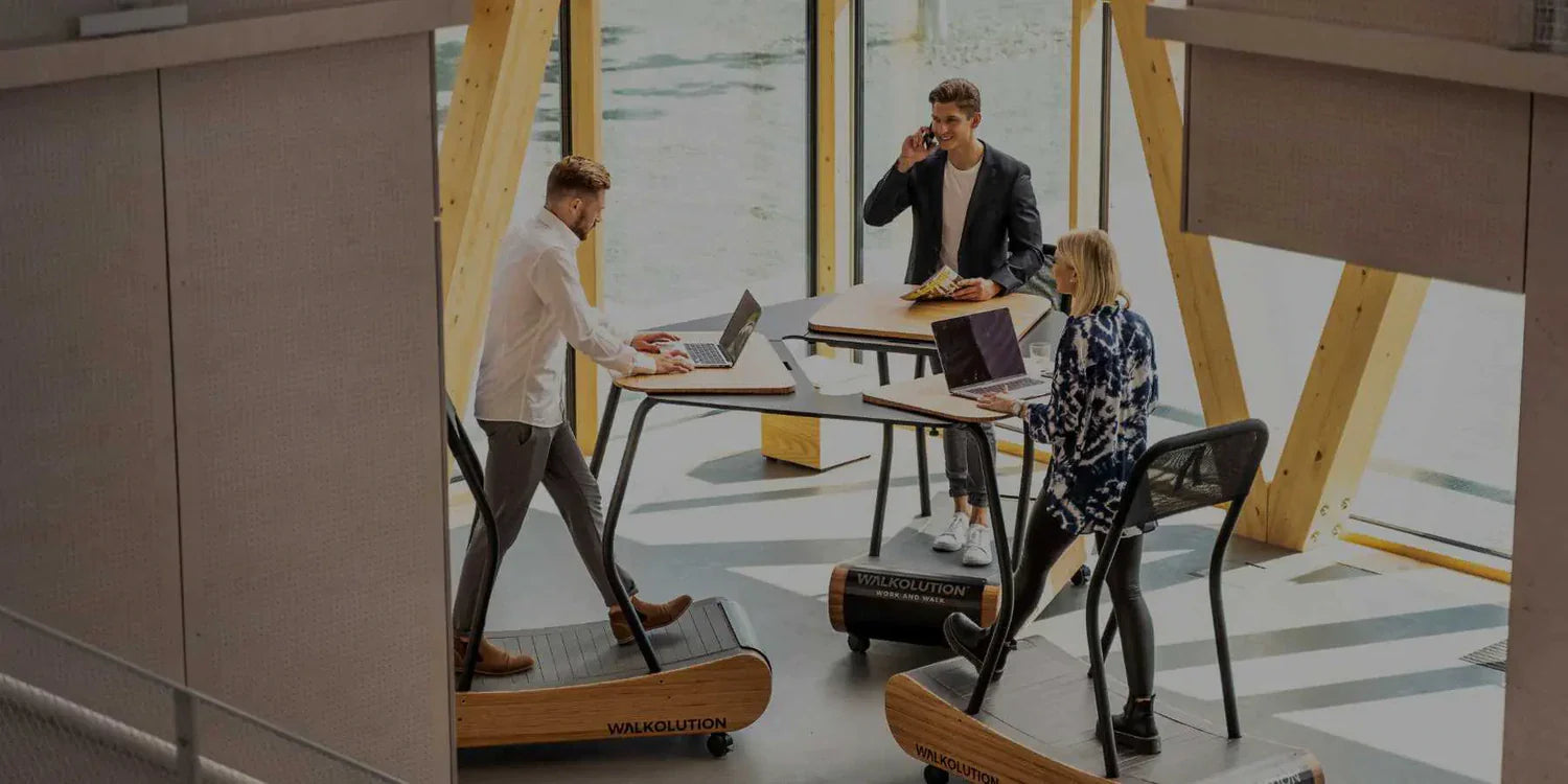 Three people work at standing desks with built-in treadmills in a modern, bright office. Two are using laptops while walking, and one is talking on the phone, all near large windows.