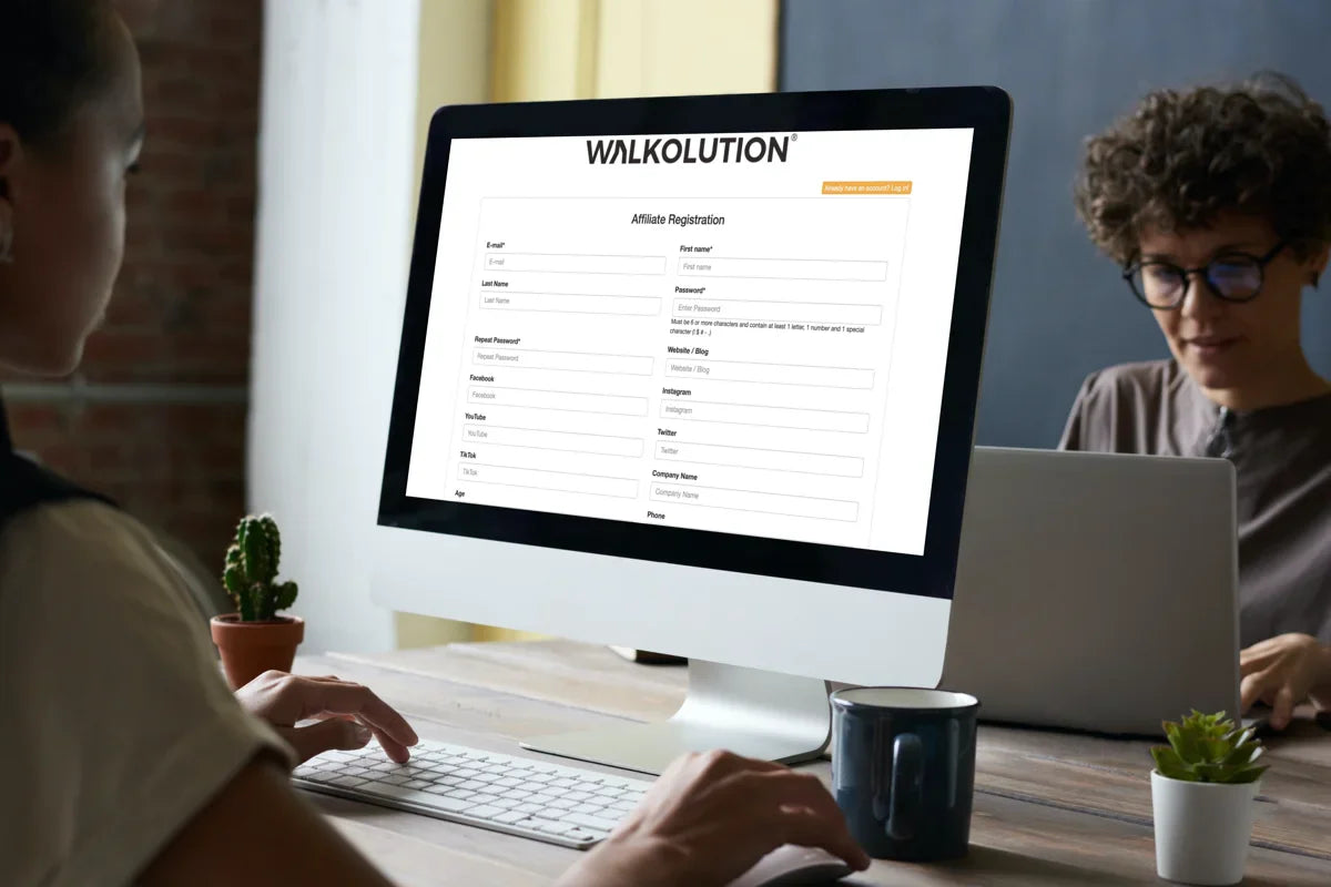 Two people work at a desk; one is using a desktop computer displaying a Walkolution affiliate registration form, while the other types on a laptop. Small plants and a cup are on the desk.