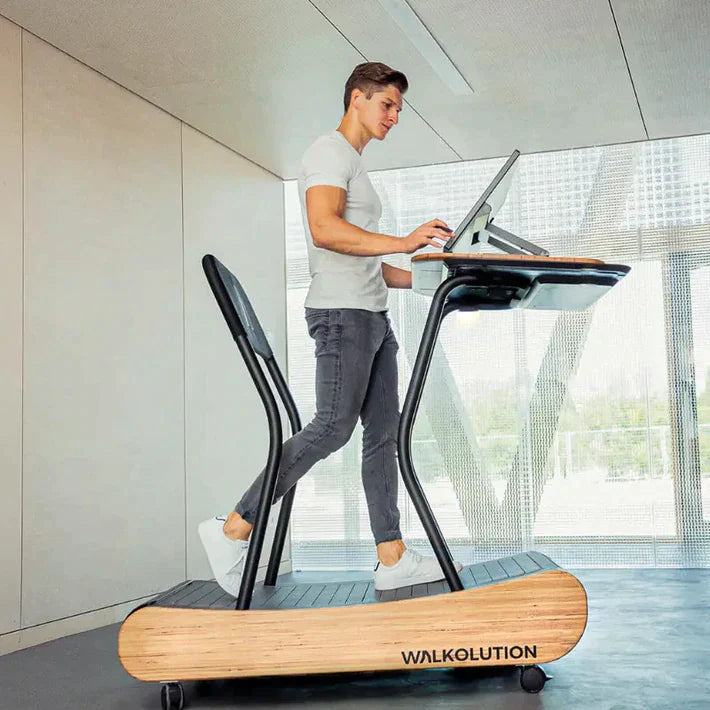 A man walks on a wooden treadmill desk, typing on a computer in a modern, bright room. The treadmill is branded WALKOLUTION and features a built-in desk and supportive backrest.