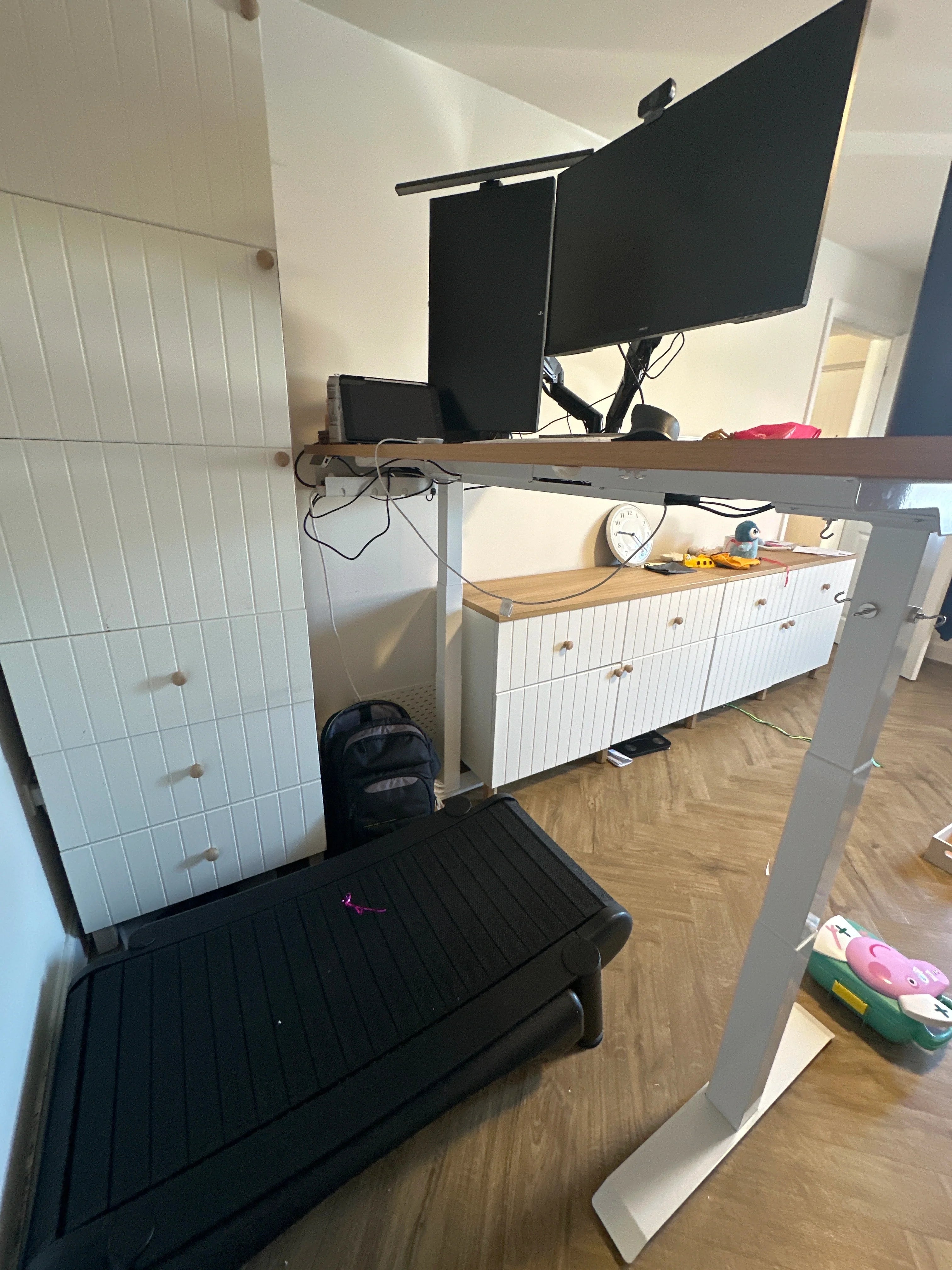 A standing desk with dual monitors and a treadmill beneath it. The setup is in a home office with cabinets, a clock, and various items, including a backpack and a childs toy, visible in the background.