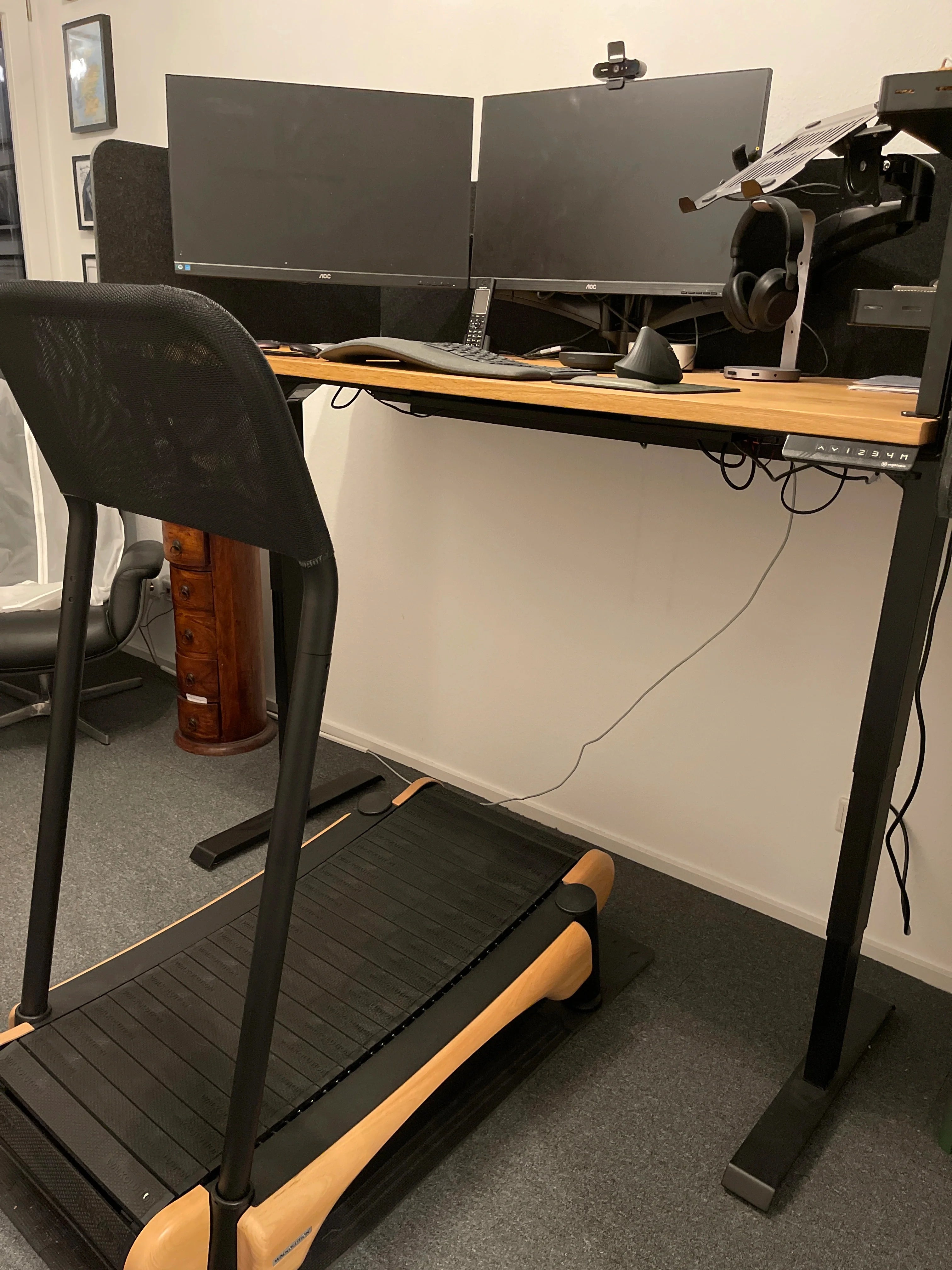A treadmill is positioned in front of a standing desk with two large monitors, a keyboard, mouse, and papers. Wires hang from the desk, and a headset is attached to the side. The room has carpeted flooring.