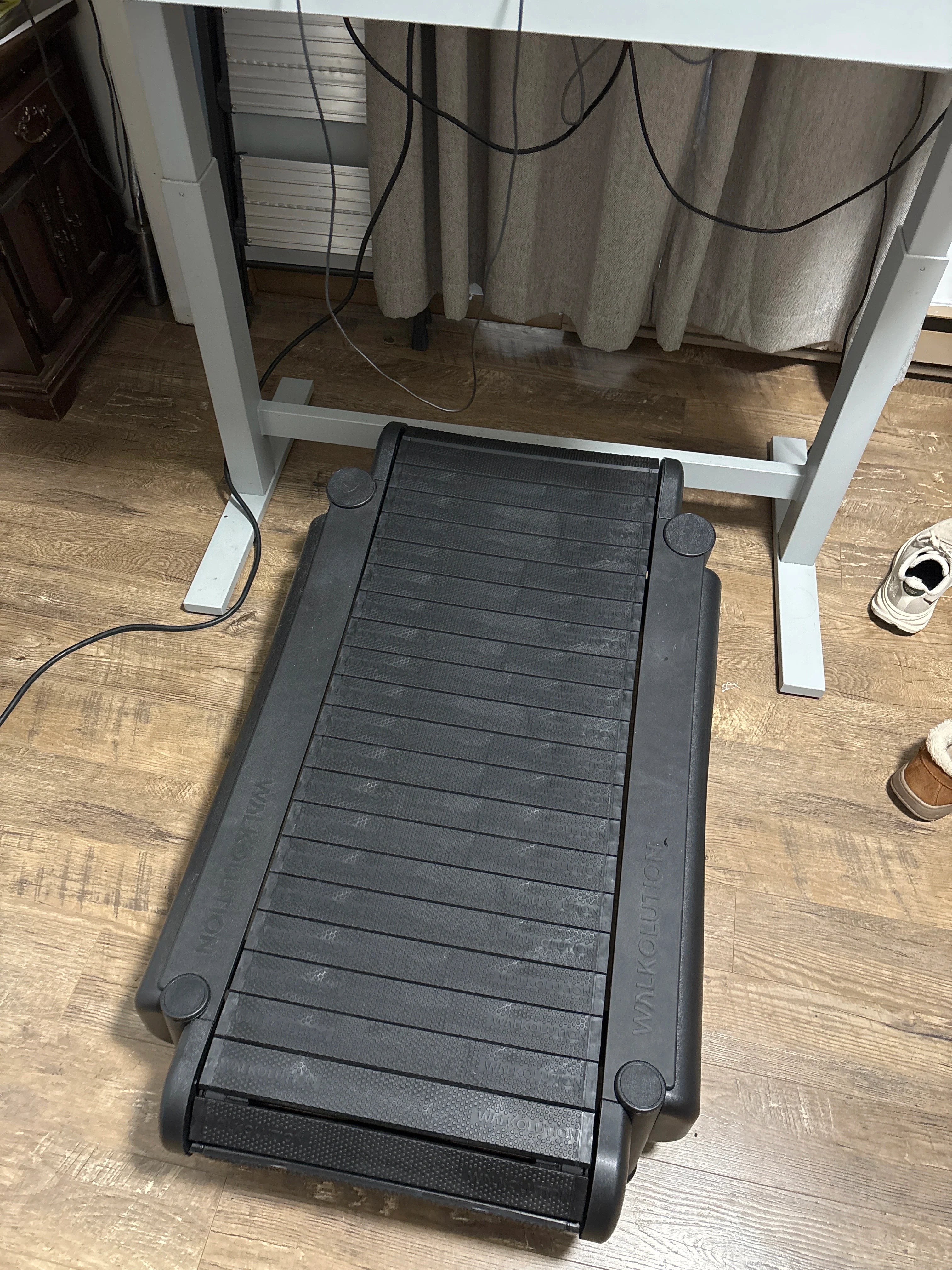 A black walking treadmill is placed under a height-adjustable desk in a room with wooden flooring. Shoes are visible to the right, and cords hang down from the desk above the treadmill.