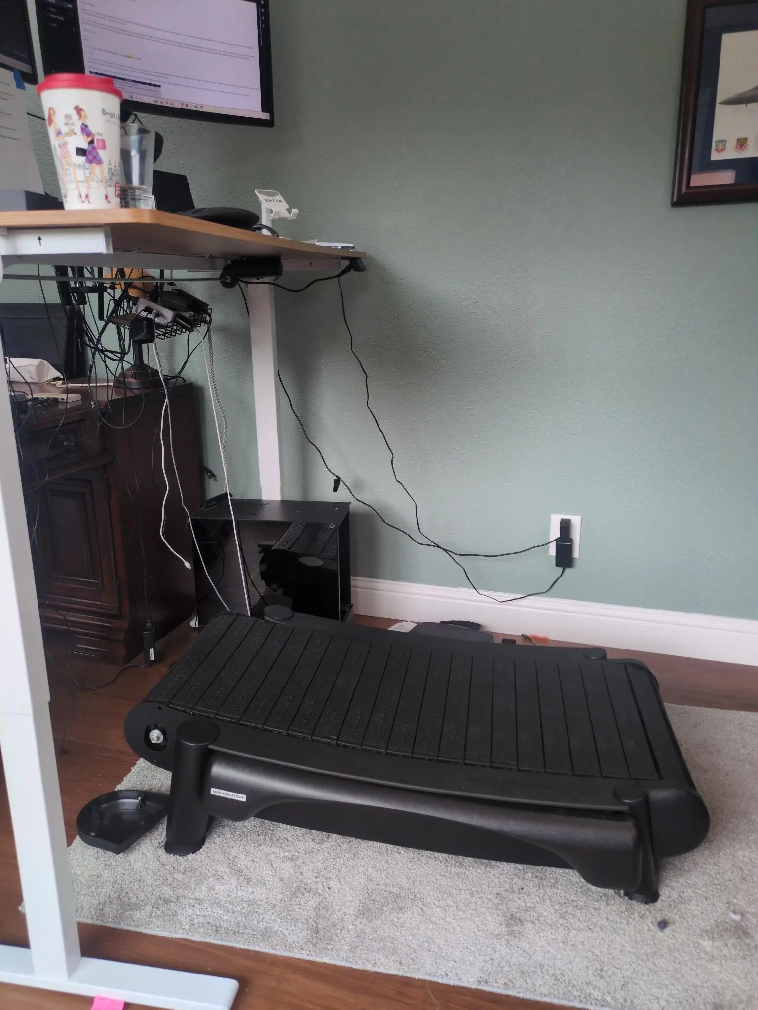 A walking treadmill is positioned under a height-adjustable standing desk, with a computer monitor and accessories on top. Wires dangle underneath, and a computer tower sits beside the desk. A travel mug is also on the desk.